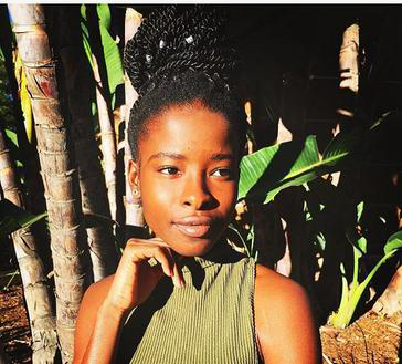 Youth Poet Laureate Amanda Gorman, a young Black woman with braids in a large updo and silver hoop earrings and a green halter shirt. She is standing with her hand under her chin in front of several palm trees. 