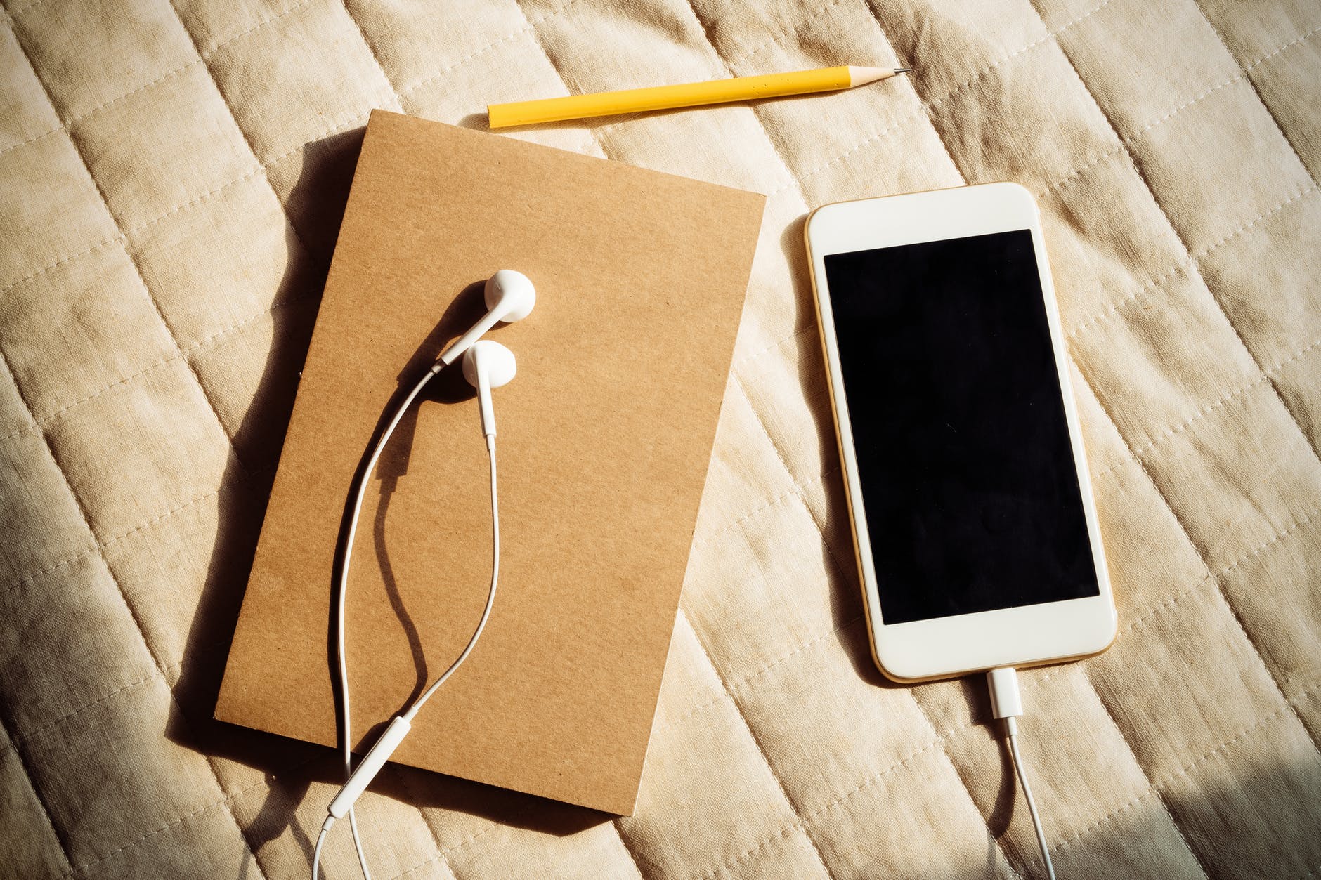 A brown cardboard notebook with white earphones connected to an Iphone with a pencil next to it on top of a bed sheet. 