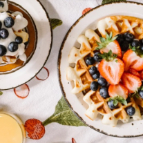 Golden brown waffles covered in blueberries and strawberry slices on white places atop a white tablecloth. 