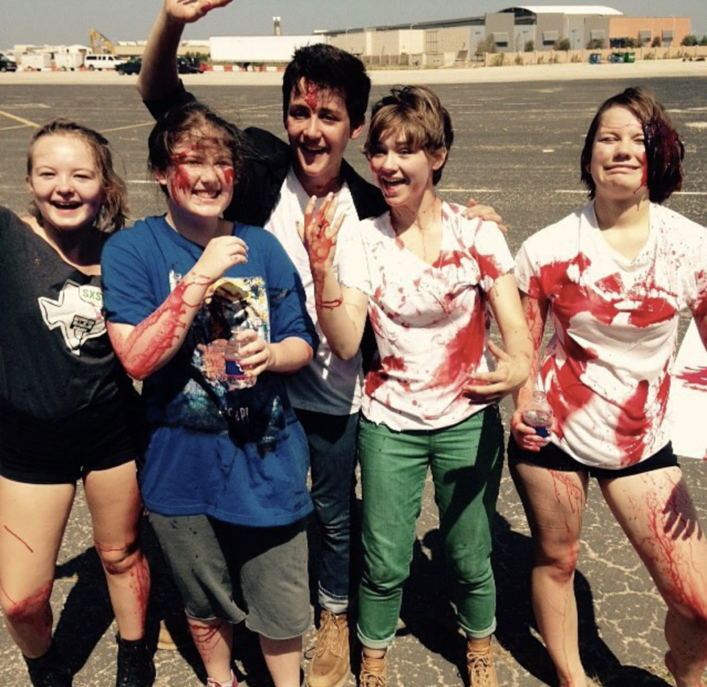 Image is of students standing outside in a group with red splotches (fake blood) splattered on them.