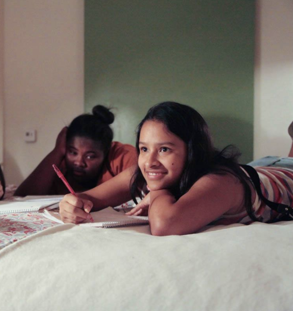 Two girls laying on a bed and one is writing as the other looks on.