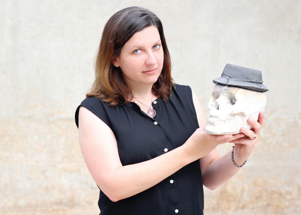 A woman with light skin and straight brown hair in a black shirt looks at the camera. She holds a white ceramic skull with a black hat.