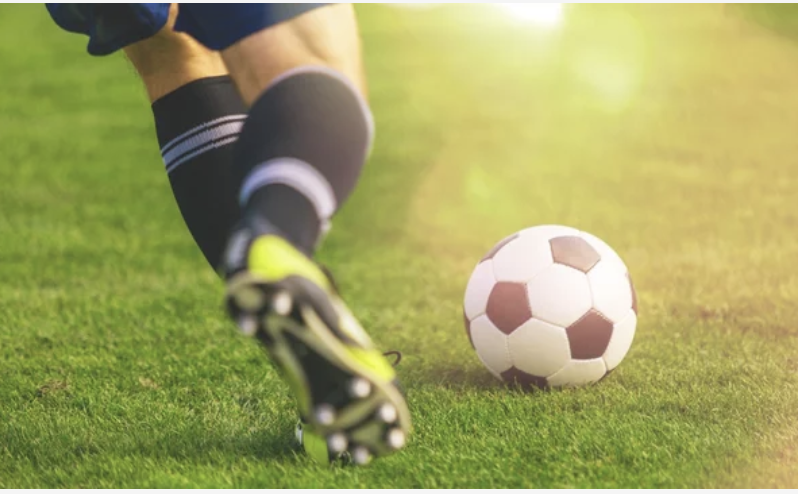 A person running and kicking a soccer ball on a bright green soccer field. They are wearing black socks and green tennis shoes with cleats.   