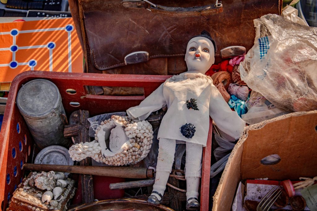 A white ceramic clown doll on top of a red crate surrounded by other old objects like a brown leather briefcase, seashells and other yard sale items. 