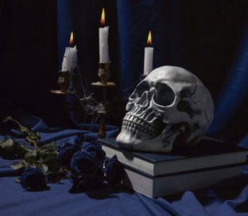 A spooky photo-realistic drawing of a skull on top of two books, with blue roses and candles in the background.