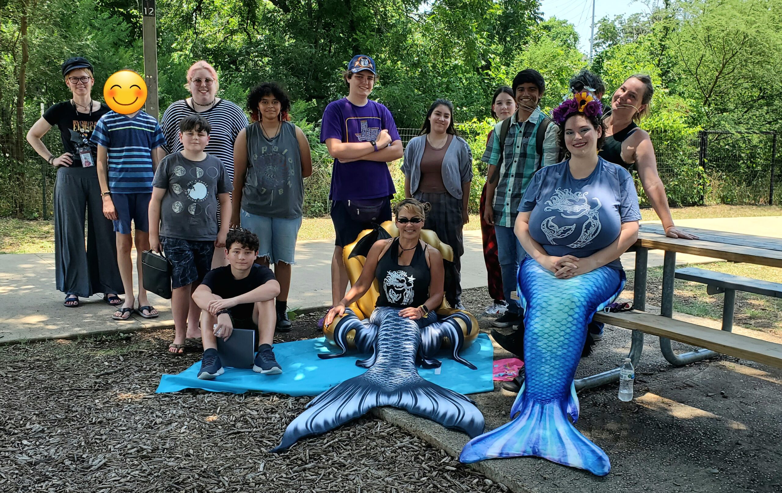 Students from the Creatures and Cryptids Camp at the San Marcos Public Library meet members of the Mermaid Society of San Marcos in a park while the mermaids in their tails pose on a bench and on the ground,