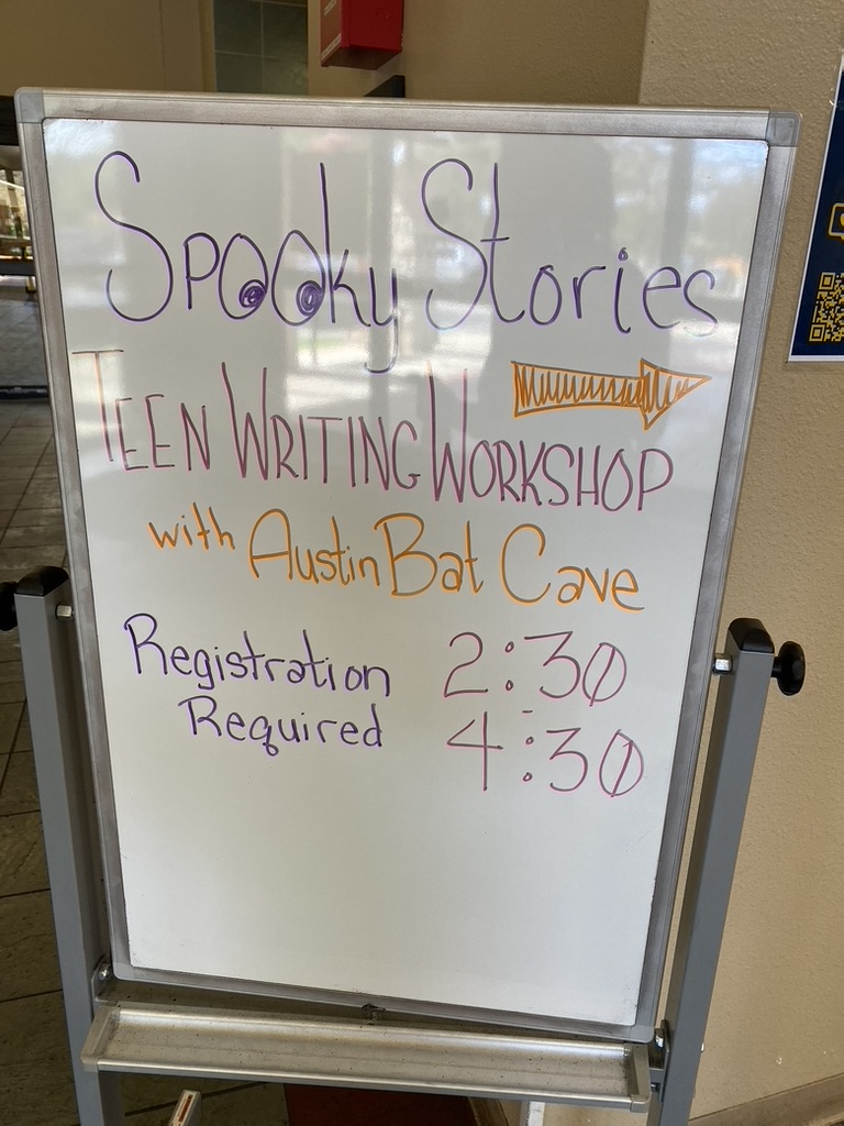 A white bulletin board with purple and orange writing that says "Spooky Stories Teen Writing Workshop with Austin Bat Cave from 2:30-4:30pm Registration Required." 