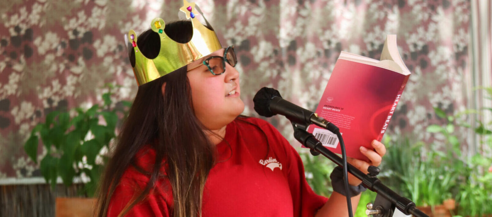 Austin Bat Cave student in a red shirt and gold crown reading her work at a microphone from the Austin Bat Cave Anthology