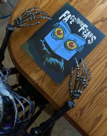 The Face Your Fears anthology on a wooden table held by black skeleton hands. The cover shows a dark blue cover with a light blue book and yellow blood shot eyes from staying up to read.