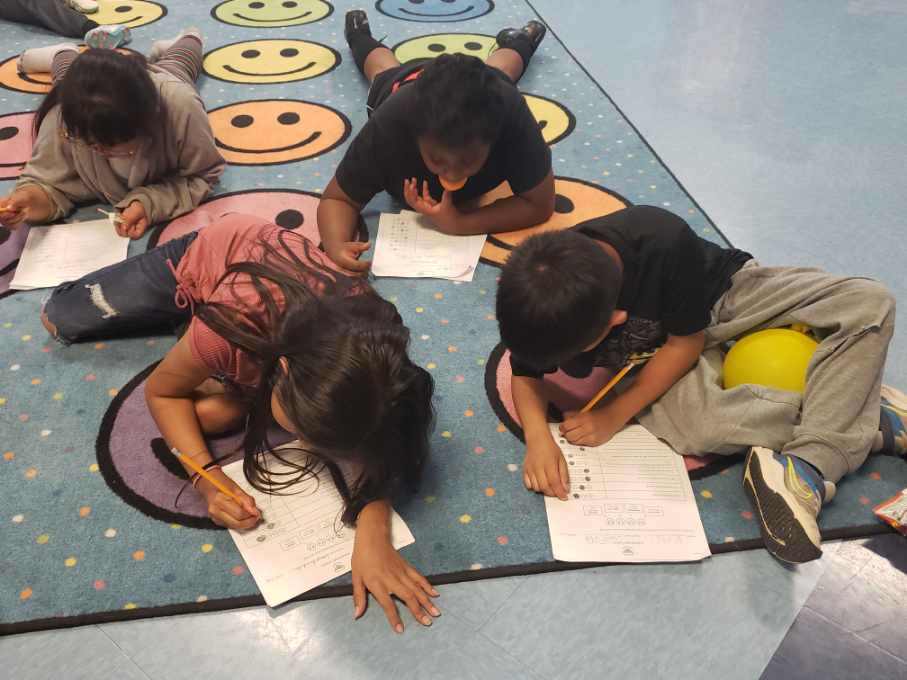 Four elementary students lying on a school classroom rug as they write in their notebooks. 