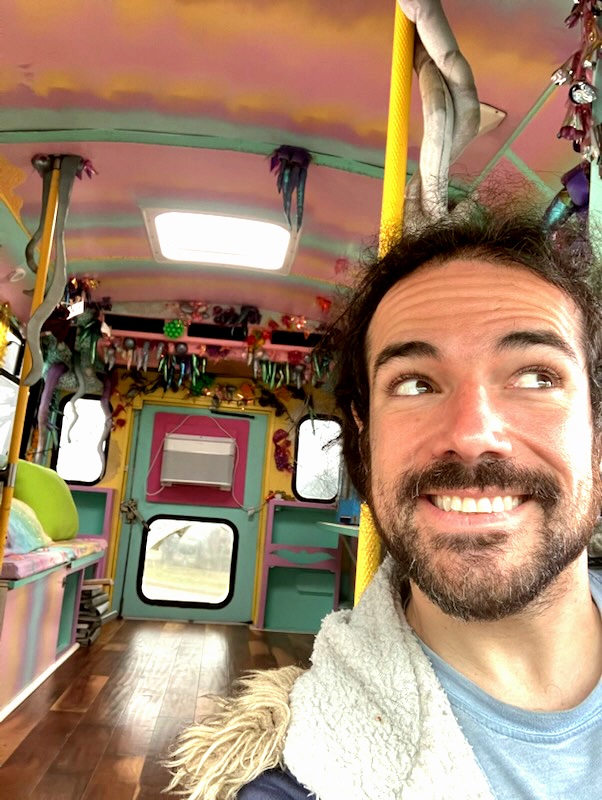 Gustavo, a light skinned person with dark brown curly long hair in a ponytail and a beard smiles at the camera in the driver's seat of the Bat Mobile bus. 