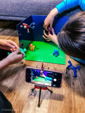 Two children manipulate toy figurines in a tiny soundstage for a stop motion film using a phone as a camera